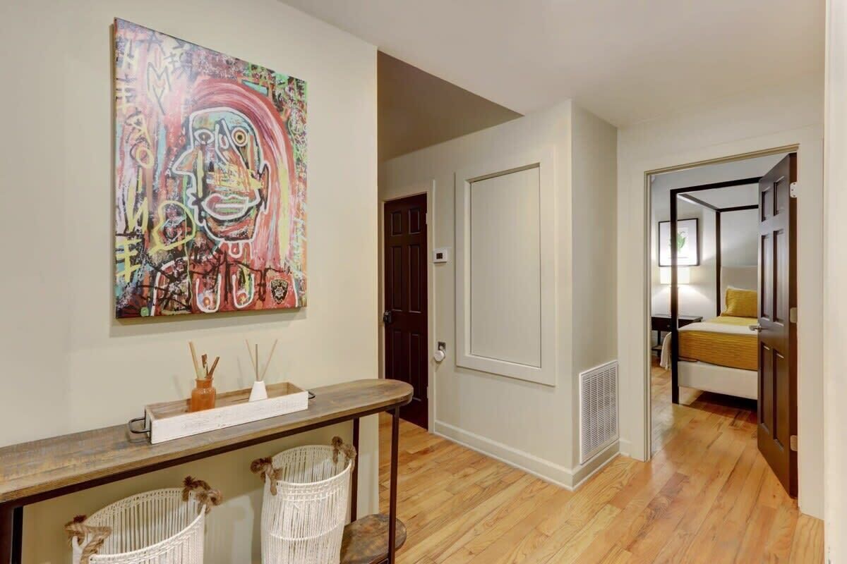 Un couloir avec un plancher en bois, une porte blanche, un lit avec un oreiller jaune et une peinture abstraite aux couleurs vives accrochée au mur.