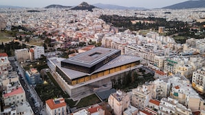 A modern building with a flat roof and large windows, surrounded by a dense urban area with various buildings and a mountain in the background. - Athenian Hillside Residence (Athens)