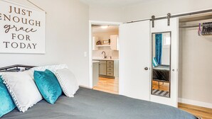 Un dormitorio con una cama, una puerta corredera hacia un baño y una cocina visible a través de una puerta abierta.