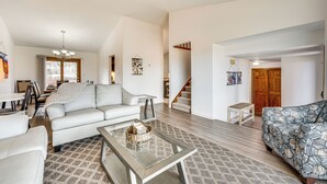 Een ruime woonkamer met een witte bank, een gestreept sofa en een salontafel. Een trap leidt naar een hoger niveau.