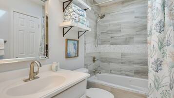A bathroom with a sink, toilet, and shower. There are shelves above the sink with towels and a framed picture on the wall.