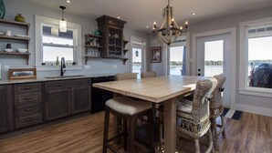 A modern kitchen with dark wood cabinets, a large island, wicker chairs, and a view of the outdoors through large windows. - Luxury Oceanfront Home in Lunenburg (First South)