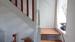 Un escalier avec des marches en bois et une rampe blanche, menant à un palier doté d’une petite fenêtre et d’un radiateur.