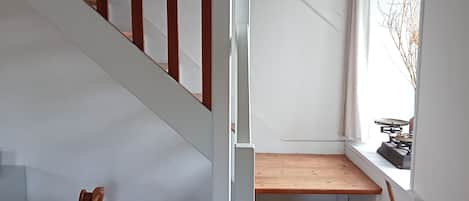 Una escalera con escalones de madera y barandilla blanca, que conduce a un rellano con un ventanal pequeño y un radiador.