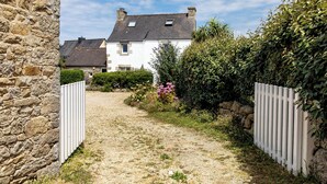 Un chemin de gravier mène à une maison blanche, bordée d’un côté par un mur de pierres et de l’autre par une clôture blanche.
