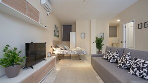 En moderne stue med et fladskærms-tv, en sofa med mønstrede puder, en seng med gavl og en potteplante.