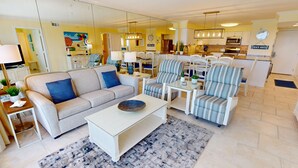 A modern living room with a beige sofa, striped armchairs, a white coffee table, and a dining area with white chairs and a striped tablecloth. - Beach House 601d 2 Bedroom Condo by RedAwning (Miramar Beach)
