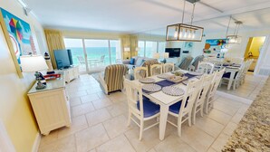 A spacious living area with a dining table set for four, a television, and a large window offering a view of the sea. - Beach House 601d 2 Bedroom Condo by RedAwning (Miramar Beach)