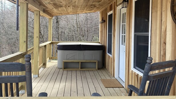 Una terraza de madera con un jacuzzi, bancos y una puerta que da a una zona cubierta.