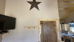 Una habitación con techo de madera, televisor montado, decoración con estrellas, puerta de madera y zona de cocina visible a través de una puerta abierta.