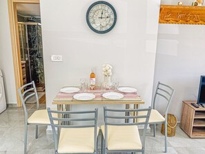 A dining area with a round wall clock, a table set with plates, glasses, and a bottle, and two chairs with cushioned seats. - Newly Built House Steps from the Beach: Everything Within Walking Distance! (Agia Marina)