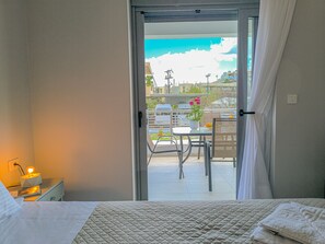 Habitación de hotel con cama, una mesita con planta y vista de la ciudad a través de una puerta abierta.