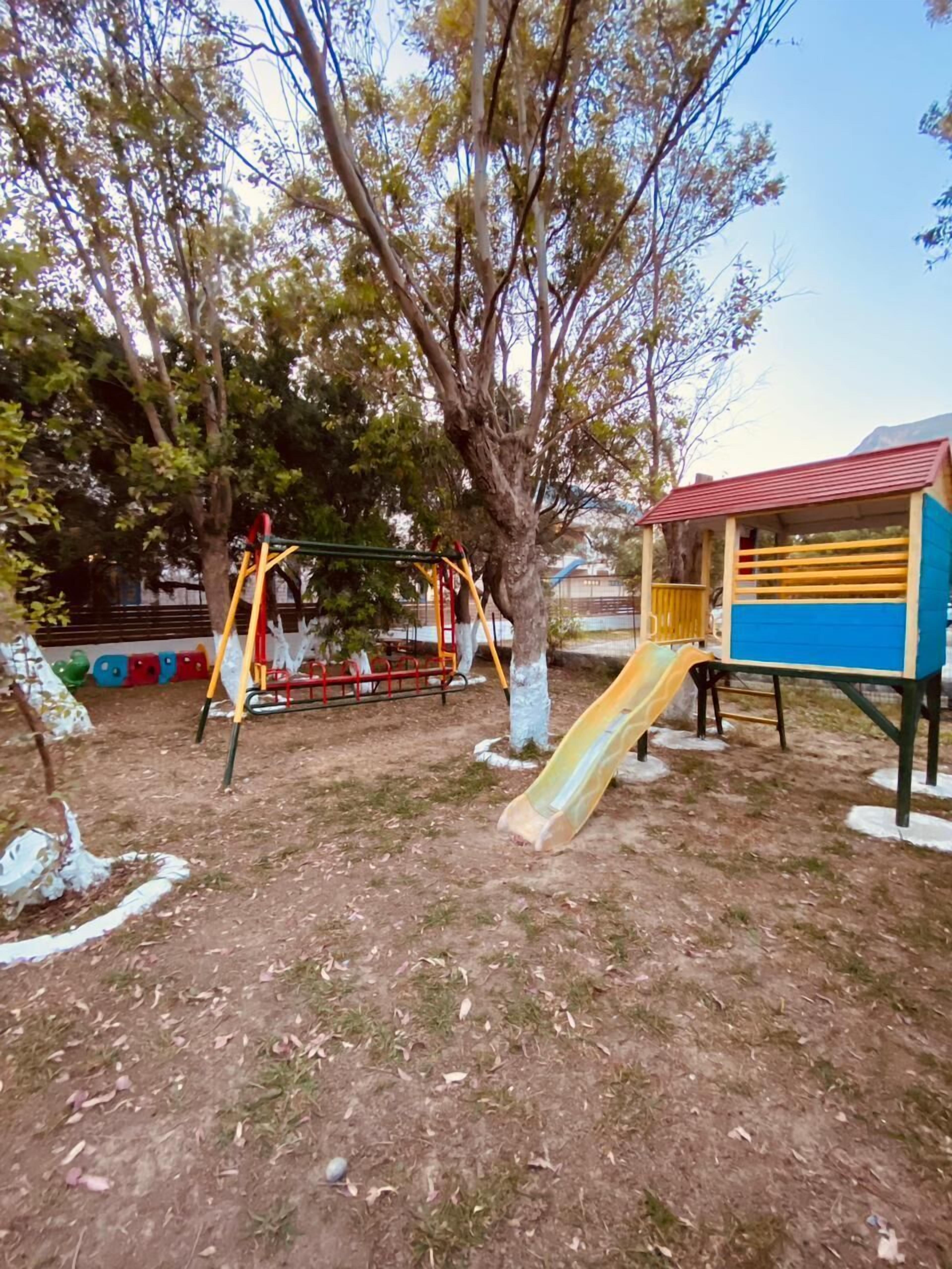 Un parque infantil con tobogán y columpios, rodeado de árboles y un pequeño edificio.