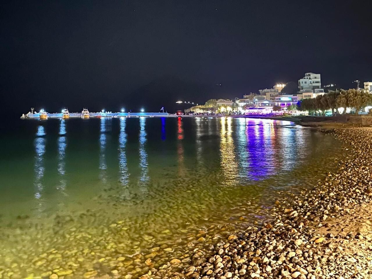 A nighttime beach scene with a pebbly shore, calm water reflecting lights, and distant buildings.