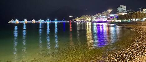 A nighttime beach scene with a pebbly shore, calm water reflecting lights, and distant buildings.
