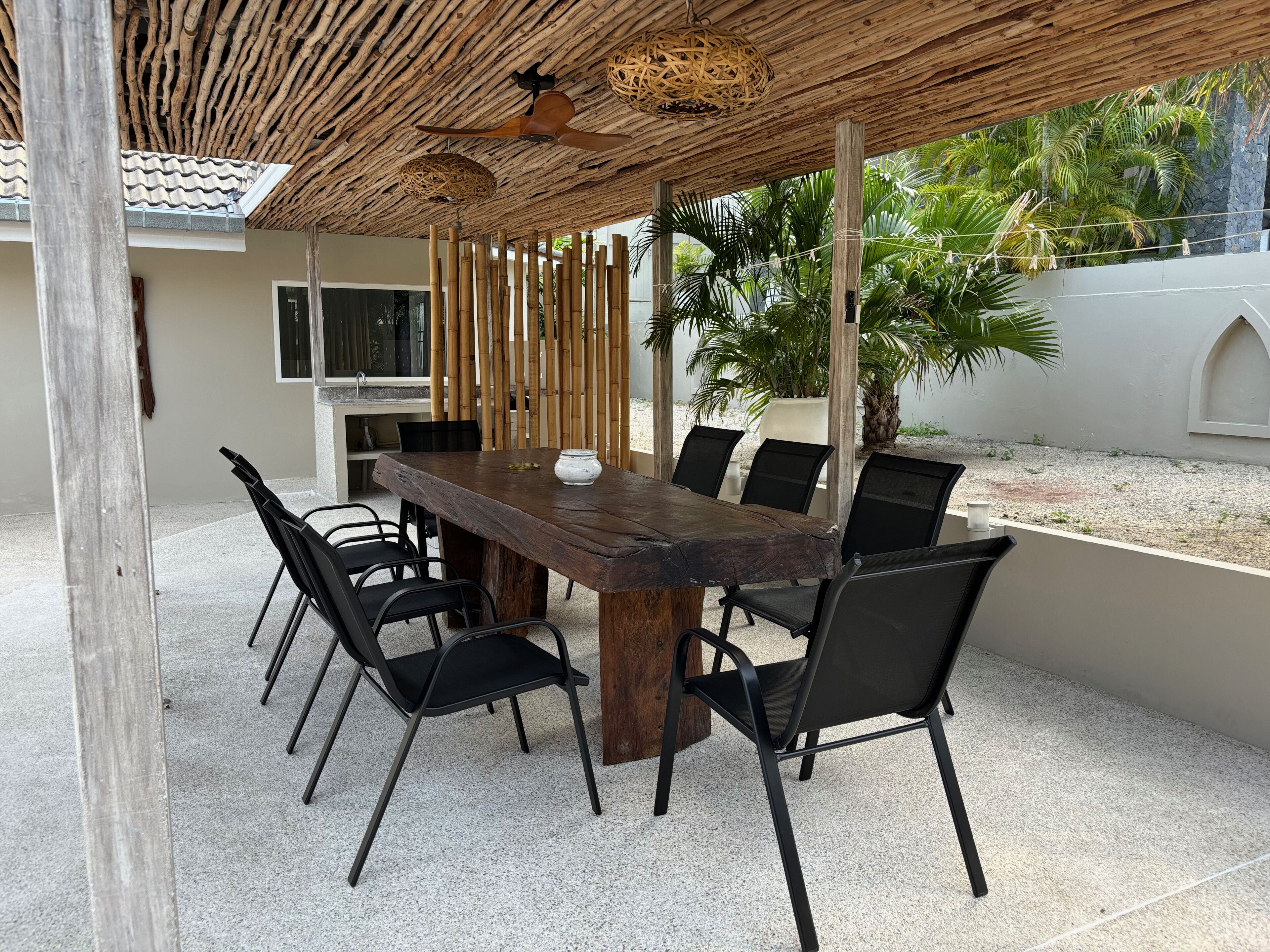 An outdoor dining area with a wooden table and chairs, a ceiling fan, and a view of a garden with palm trees.