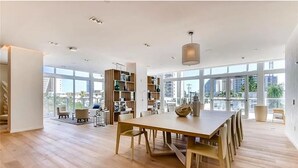 A spacious living area with a large wooden dining table, a built-in bookshelf, and a view of the city through large windows.