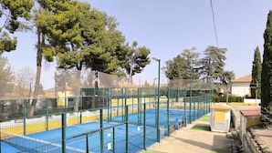 Una cancha de tenis exterior con una valla verde, rodeada de árboles y un cielo despejado.