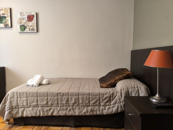 A neatly made bed with a quilted blanket, a brown pillow, and rolled towels on the side. There is a lamp on a bedside table and artwork on the wall.