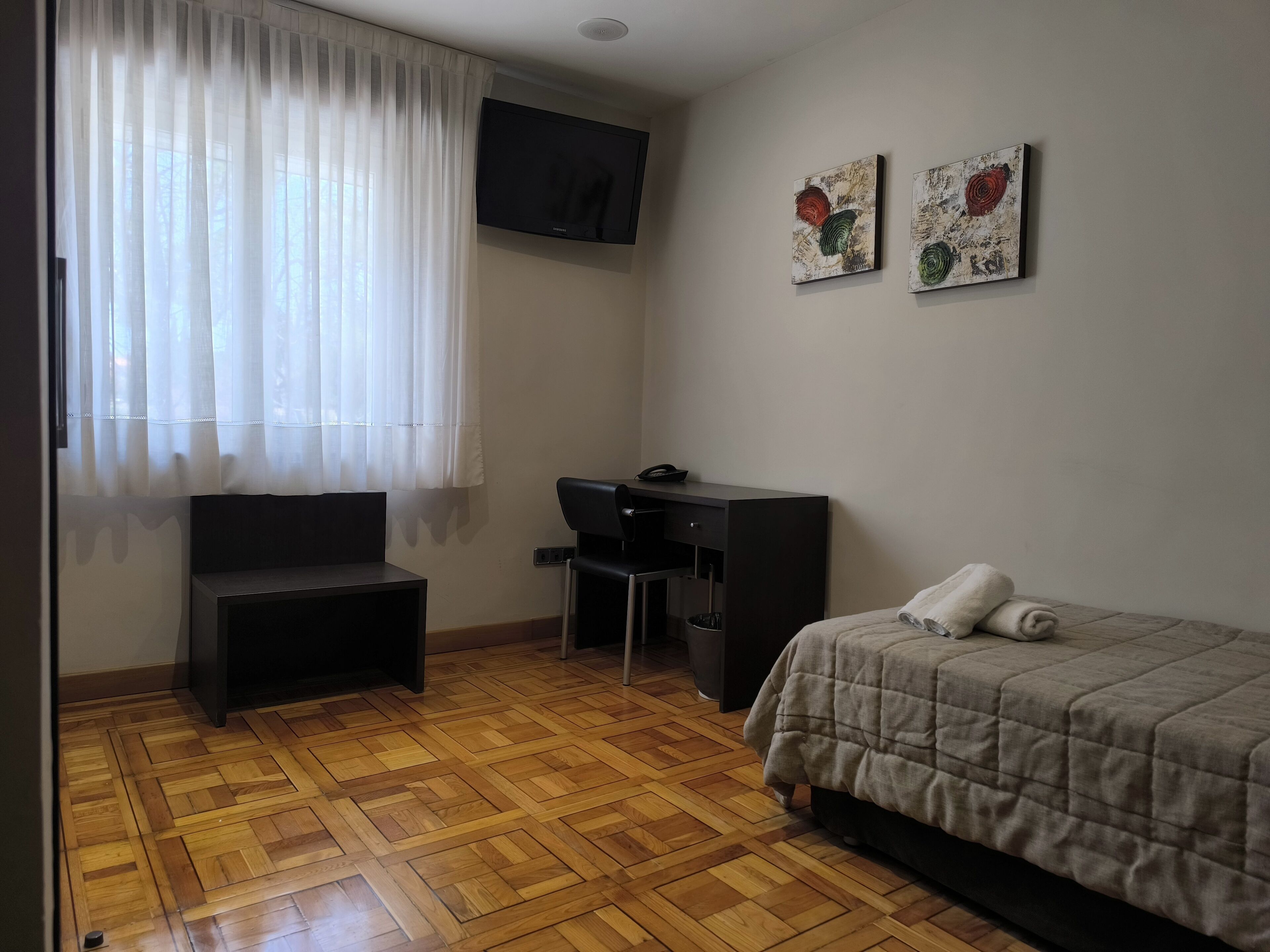 A small, neatly arranged room with a bed, a desk, a chair, and a television on the wall.