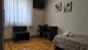 A small, neatly arranged room with a bed, a desk, a chair, and a television on the wall.