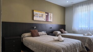 Habitación de hotel con dos camas, un cabecero oscuro, una mesita de noche pequeña, una obra de arte colgada en la pared y una ventana con cortinas translúcidas.