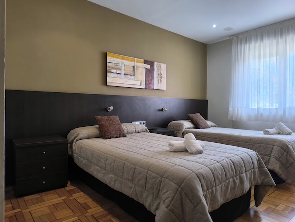 Habitación de hotel con dos camas, un cabecero oscuro, una mesita de noche pequeña, una obra de arte colgada en la pared y una ventana con cortinas translúcidas.