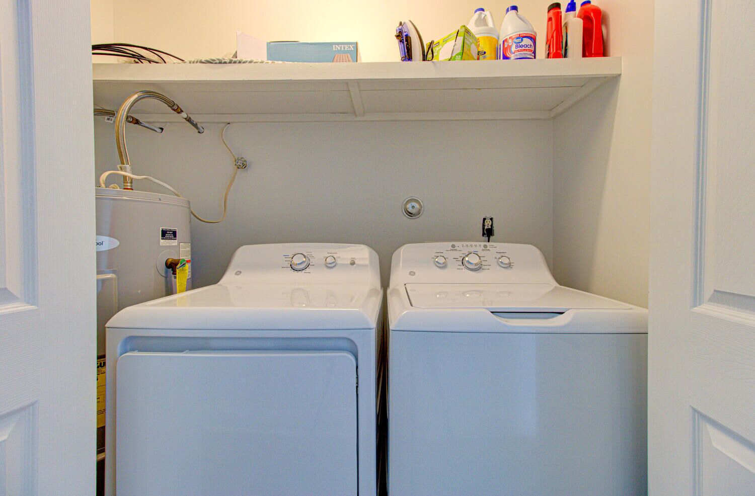 Ein Waschraum mit zwei weißen Waschmaschinen mit Frontlader, einem Warmwasserbereiter und einem Regal mit Reinigungsmitteln.