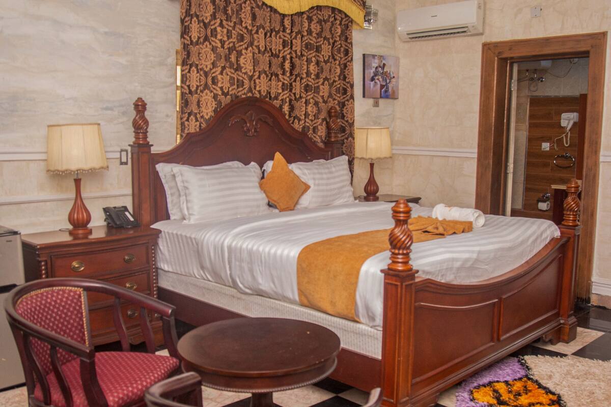 A hotel room with a large bed, two bedside tables with lamps, a wooden chair, a round table, and a checkered floor.