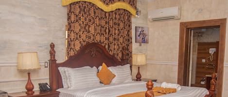 Habitación de hotel con una cama grande, dos mesitas de noche con lámparas, una silla de madera, una mesa redonda y un suelo a cuadros.