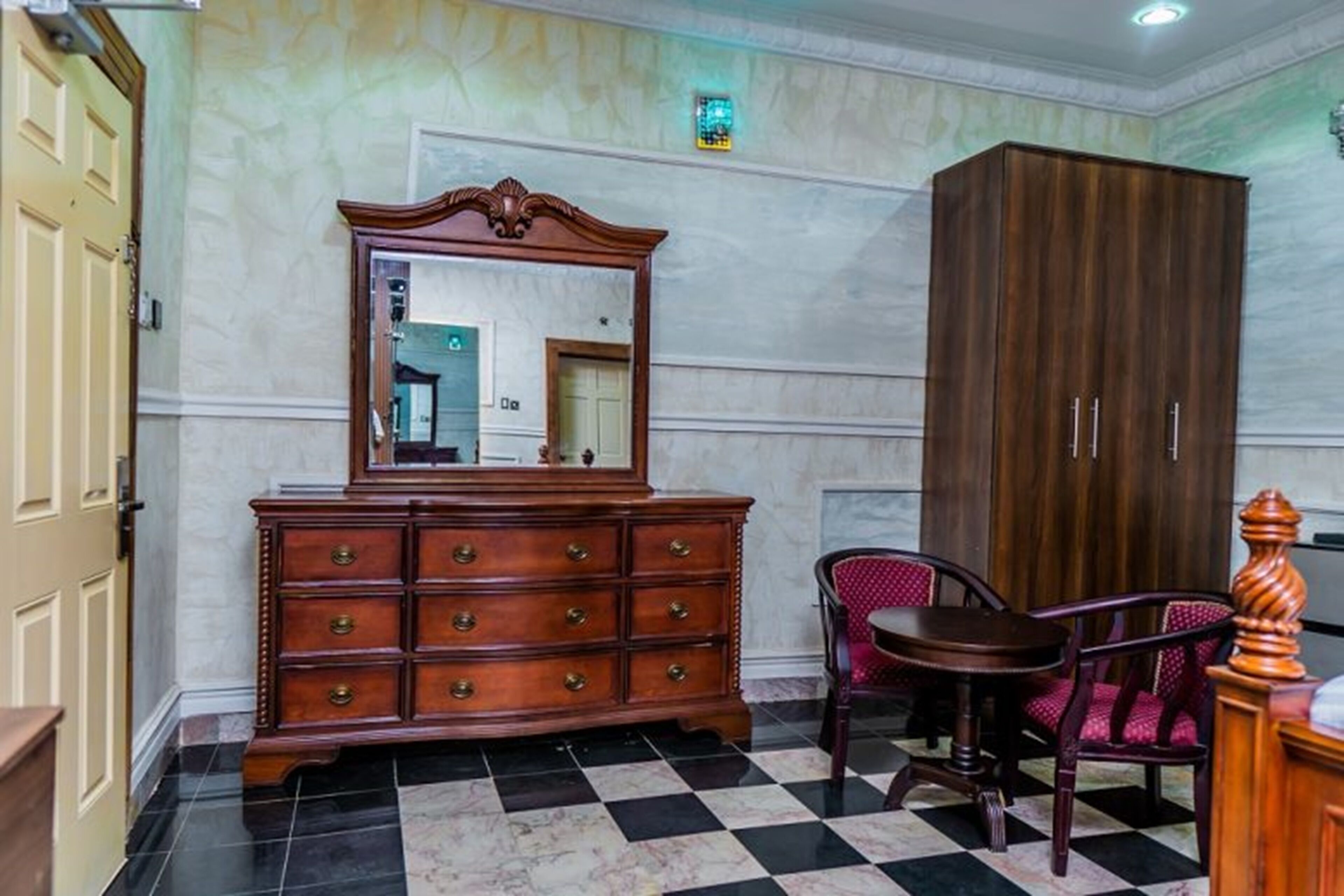A room with a checkered floor, a wooden dresser with drawers, a mirror, a red upholstered chair, and a tall wooden wardrobe.