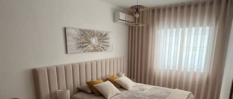 A bedroom with a bed, a nightstand, a chair, a window with curtains, and a wall art piece.