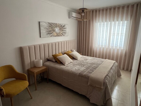 A bedroom with a bed, a nightstand, a chair, a window with curtains, and a wall art piece.