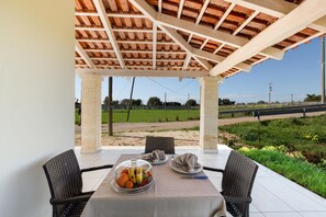 Une terrasse couverte avec une table dressée pour deux personnes, agrémentée d’un panier de fruits et d’une bouteille.
