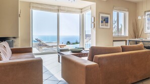 A living room with a large sliding glass door leading to a balcony with a view of the sea. There are two sofas and a small table with books and a bowl.