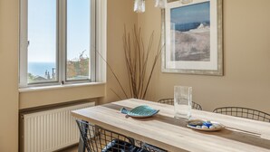 A dining area with a table, chairs, a view through a window, and a framed painting on the wall.