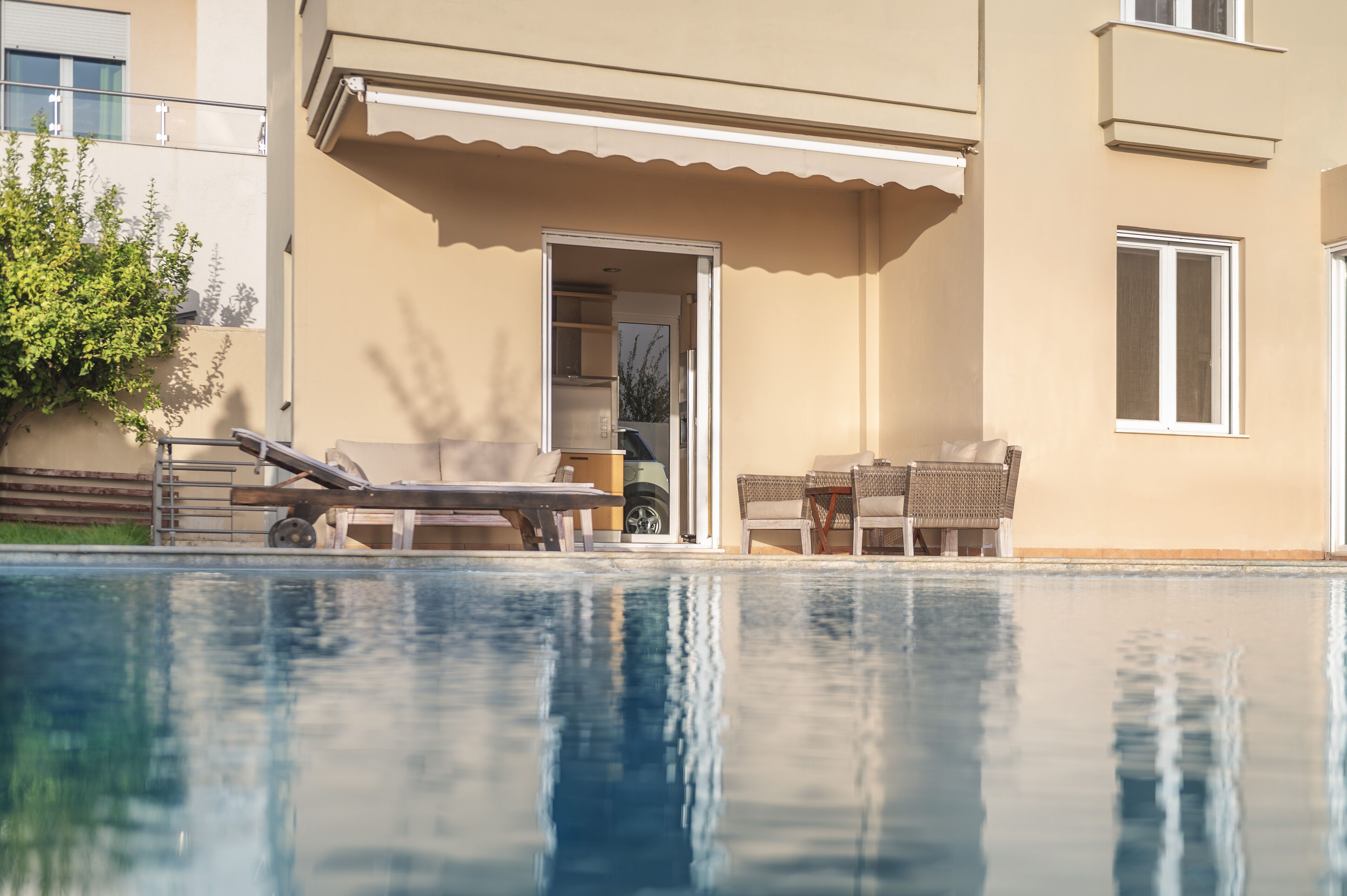 Une piscine avec des chaises longues et une table, un bâtiment avec une porte et des fenêtres, et un arbre en arrière-plan.