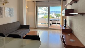 A modern hotel room with a sofa, a small table, a TV, a balcony with a view of buildings and mountains, and a wall-mounted shelf. - Agi Port Canigo 206 Apartamento for rent in Roses Girona. (Roses)