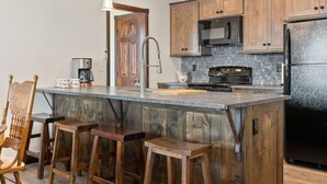 A kitchen with wooden cabinets, a large island with stools, a stainless steel refrigerator, and a coffee maker. - Family Friendly Cabins w/ Mountain & Wildlife View (Kalispell)