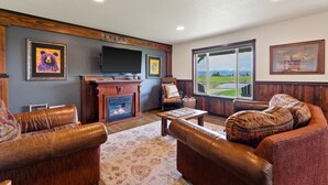 A cozy living room with leather sofas, a fireplace, and large windows offering a view of a golf course. - Family Friendly Cabins w/ Mountain & Wildlife View (Kalispell)