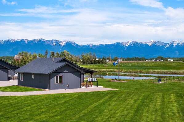 A modern building with a dark exterior, a covered patio, and a view of snow-capped mountains in the background. - Family Friendly Cabins w/ Mountain & Wildlife View (Kalispell)