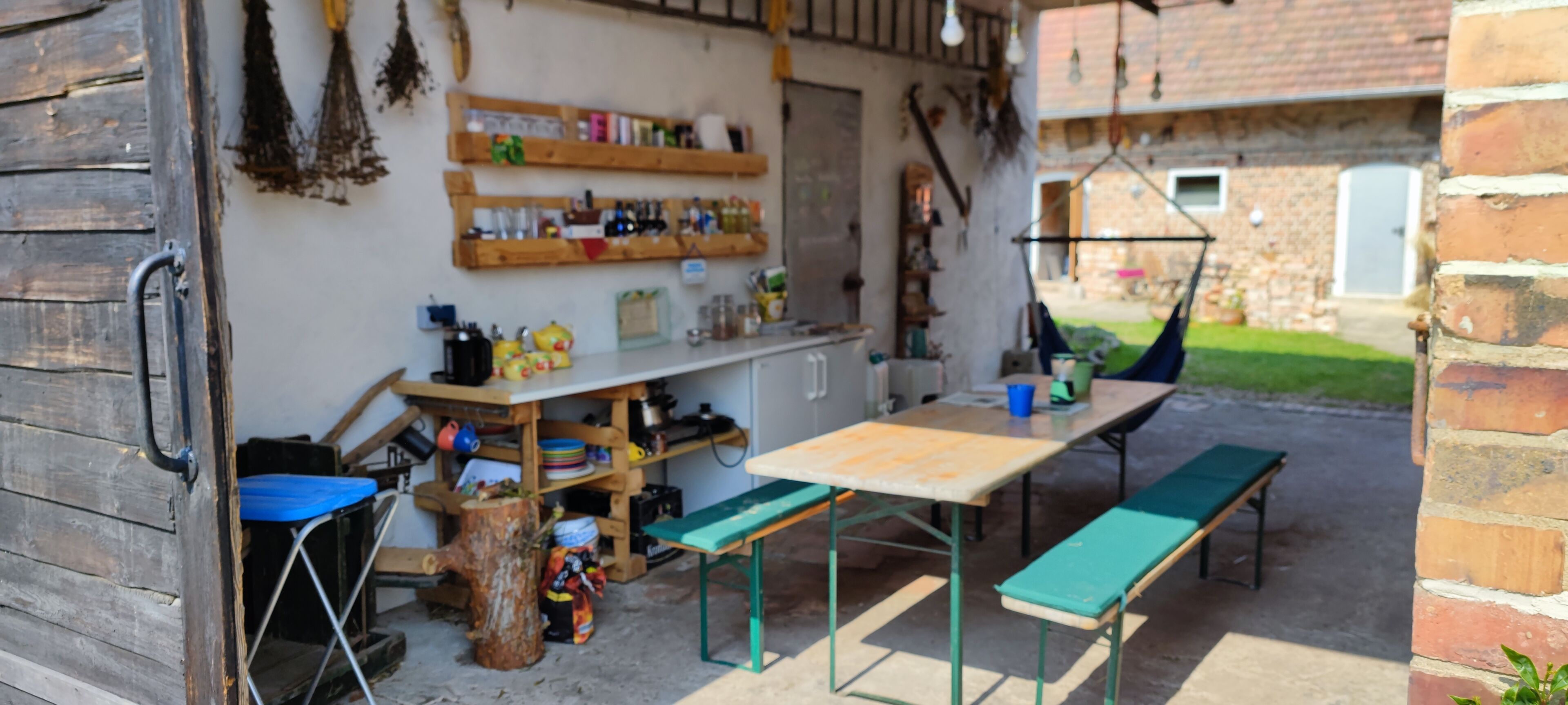 Un espace extérieur rustique aménagé avec une table et des bancs en bois, entouré d’étagères remplies de divers objets.