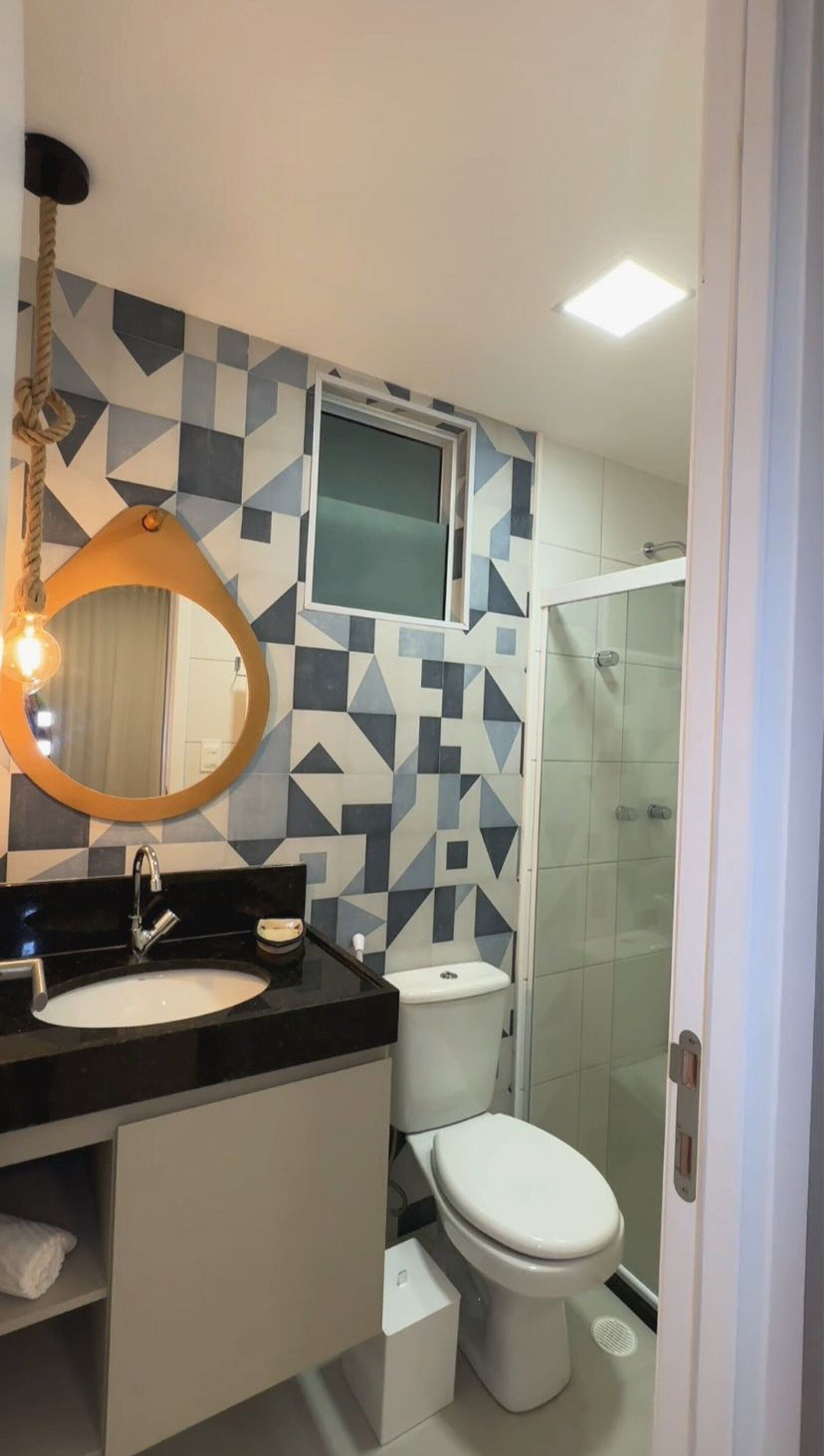 Ein modernes Badezimmer mit einem geometrischen Fliesenrückwand, einem runden Spiegel mit einer Glühbirne, einer weißen Toilette und einer Duschkabine aus Glas.