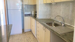 A compact kitchen with light wooden cabinets, a stainless steel sink, and a white refrigerator. - Agi Europa 62 Apartamento for rent in Roses Girona. (Roses)