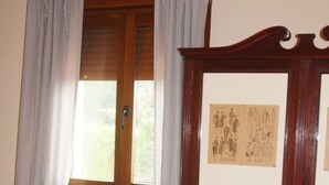 A room with a wooden cabinet with framed pictures, a radiator, a window with curtains, and a bed with a table. - Short description of the property (San Juan de Duz)