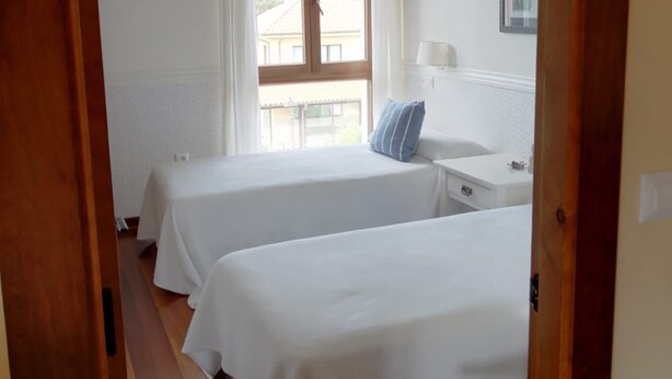A bedroom with two beds, a wooden floor, a window with curtains, and a small table with a lamp. - Short description of the property (San Juan de Duz)