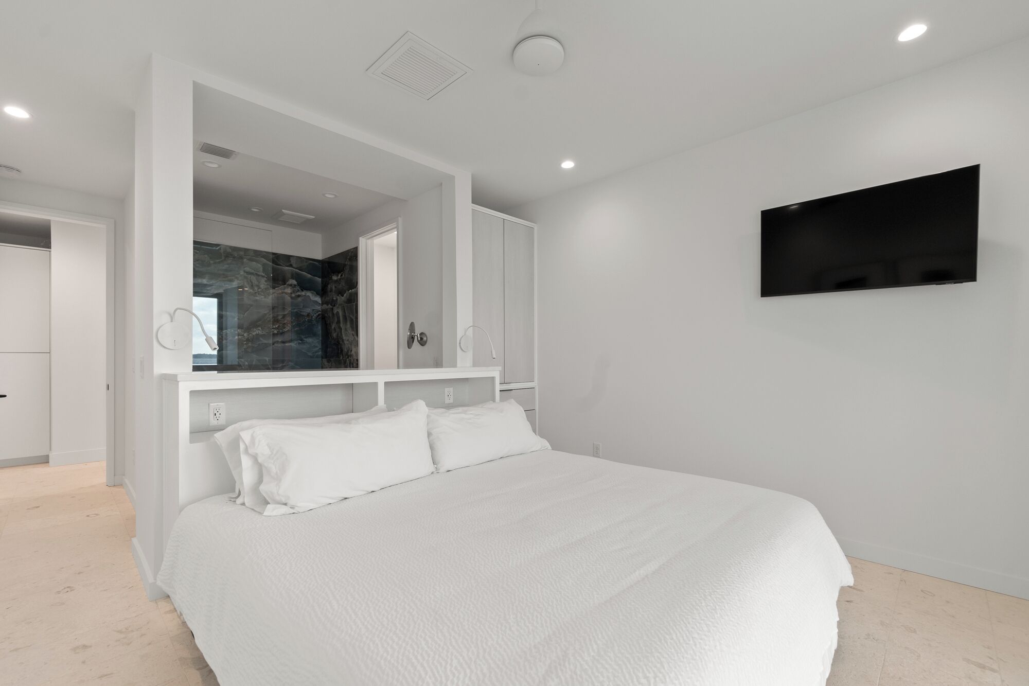 Un dormitorio moderno con una cama, un televisor montado en la pared y un baño visible a través de una puerta abierta.