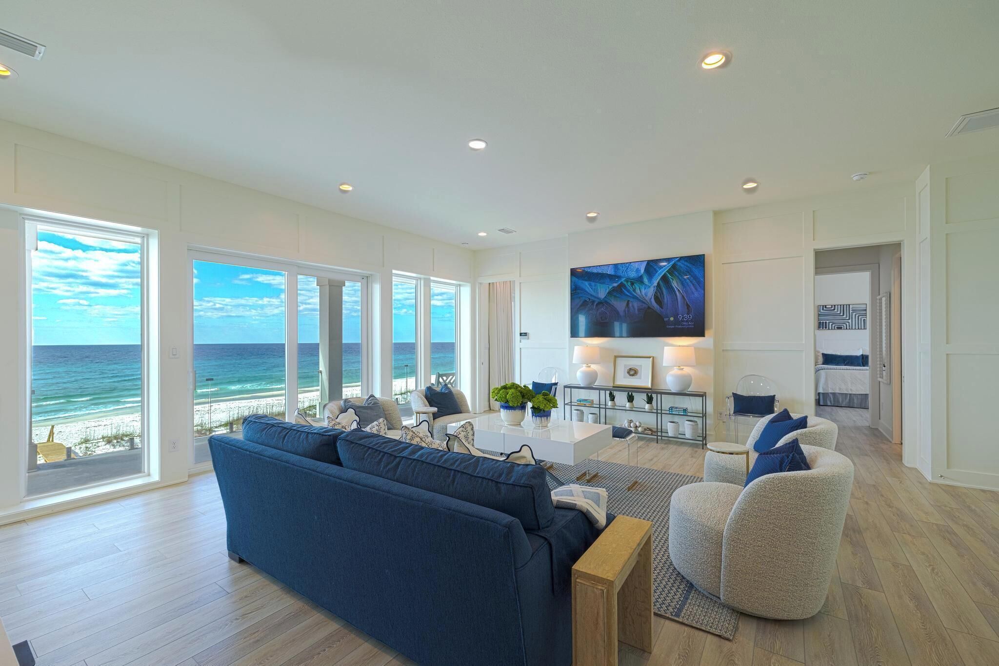 A spacious living room with a large sofa, a coffee table, and a TV mounted on the wall. The room has large windows offering a view of the ocean.