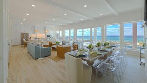 Un vaste espace de vie comprenant un coin repas avec une table pour six personnes, de grandes fenêtres offrant une vue sur la plage et une cuisine dotée de meubles blancs.