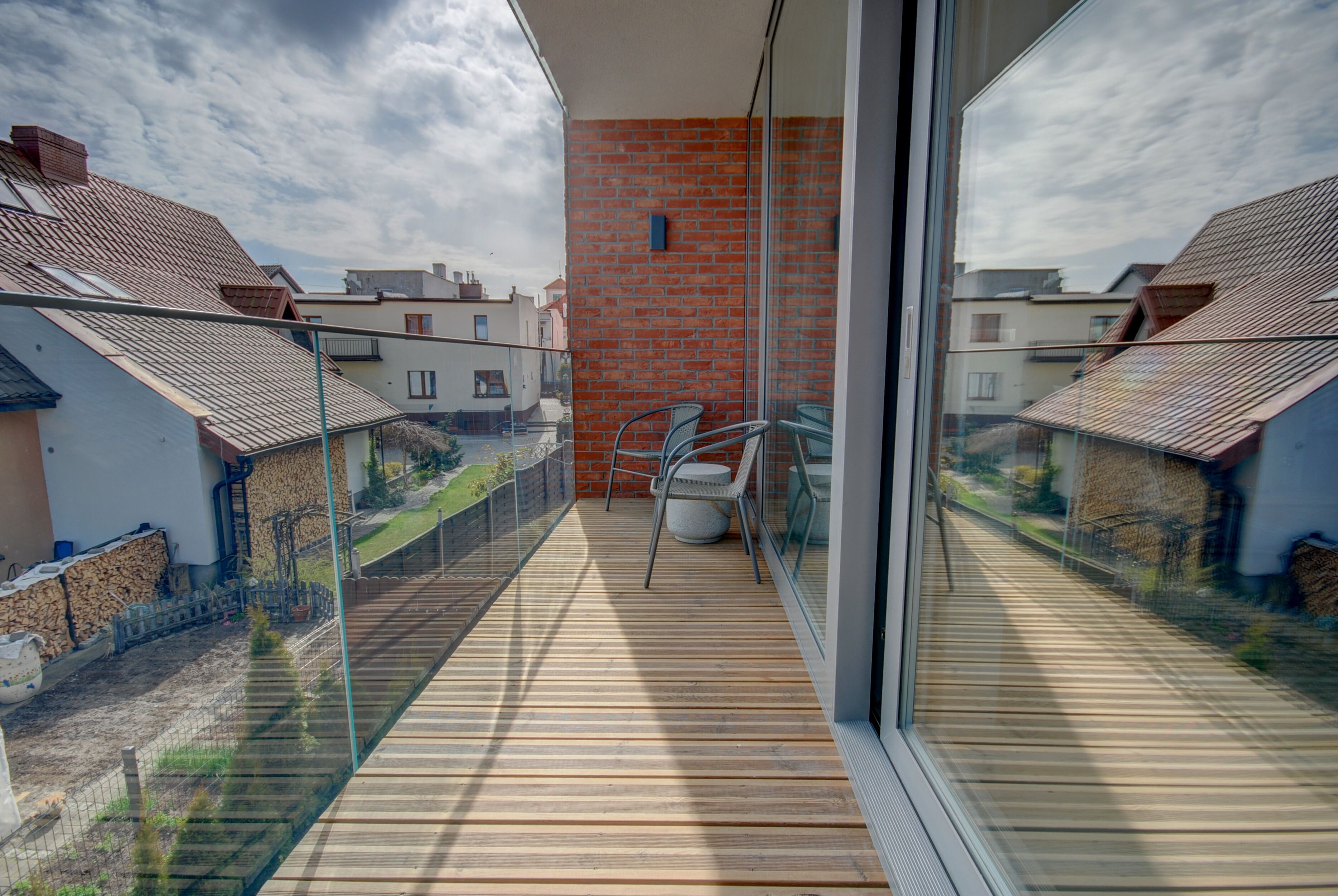Un balcone con pavimento in legno, ringhiera in vetro e vista sulle case vicine.